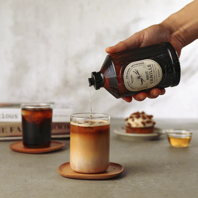 Person pouring Bacanha Vanilla from a bottle into a glass of coffee on a table with a blurred background.