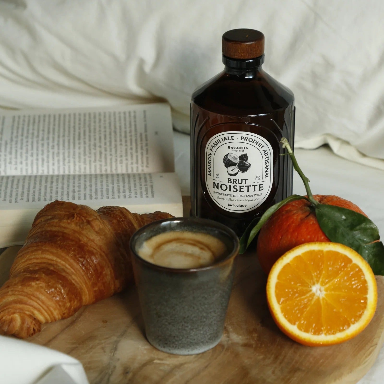 Bottle of Bacanha Brut Noisette coffee, croissant, cup of coffee, and oranges on a wooden board.