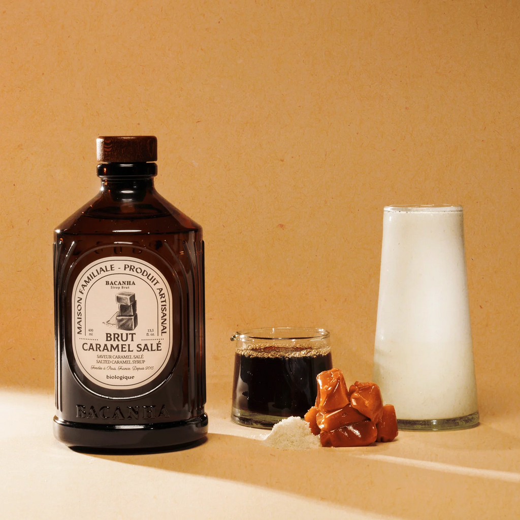 Bruine glazen bacanha gezouten karamel siroop fles op naast een hoopje zout en karamelblokjes op een lichtgele achtergrond
